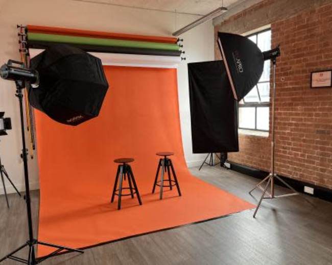 The image shows a photography studio setup with an orange backdrop, two black stools, and various lighting equipment.