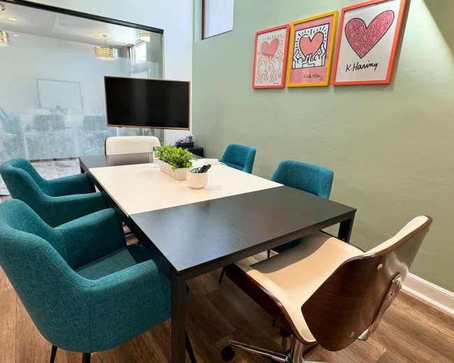 A modern meeting room with a rectangular black table surrounded by six chairs, three in teal and two in cream, alongside a television and framed heart artwork on the wall.