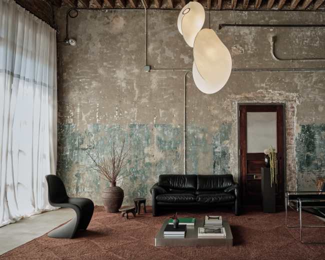 The image shows a minimalist living space with a black leather sofa, a glass coffee table, and an artistic light fixture hanging from the ceiling, set against a textured wall with peeling paint.