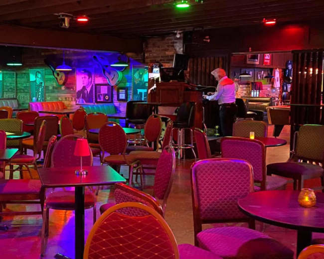 The image shows a dimly lit lounge with colorful lighting, featuring empty tables and chairs, and a lone figure attending to a stage.