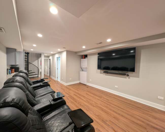 The image shows a spacious room with wooden flooring, featuring black reclining chairs facing a large wall-mounted television and a spiral staircase in the corner.