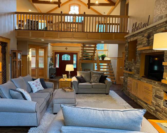 A spacious living room features comfortable gray sofas, a wooden staircase, and large windows allowing natural light to fill the space.