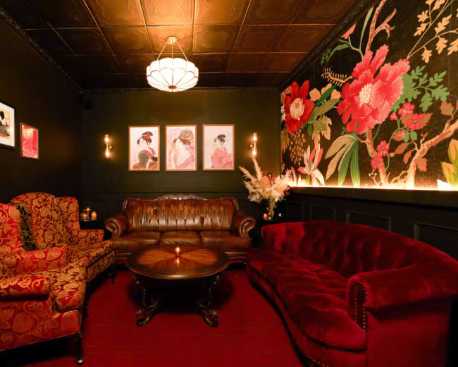 The image shows a dimly lit lounge with dark walls, featuring ornate furniture, floral wallpaper, and framed portraits.