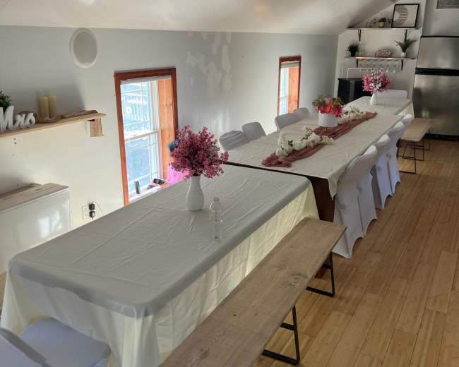 A spacious indoor area set up for a gathering, featuring two long tables covered with white tablecloths and decorated with floral arrangements, flanked by chairs along the sides.