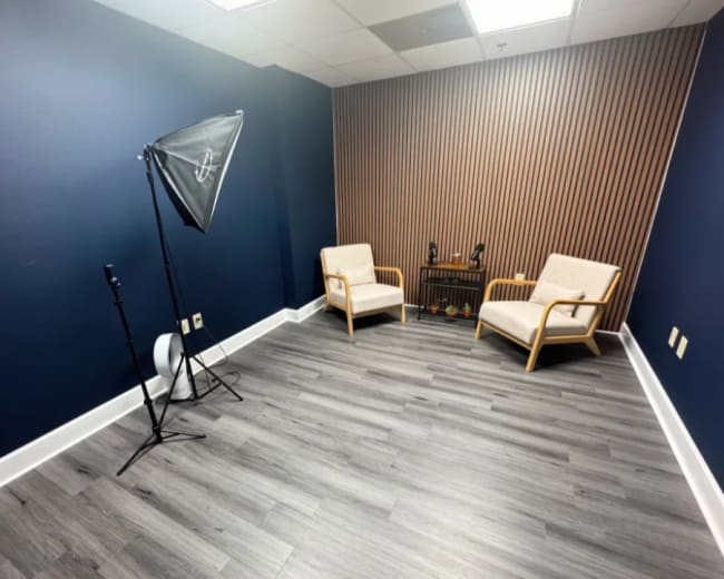 The image shows a small, modern room with two chairs, a small table, and a backdrop featuring vertical wooden slats.
