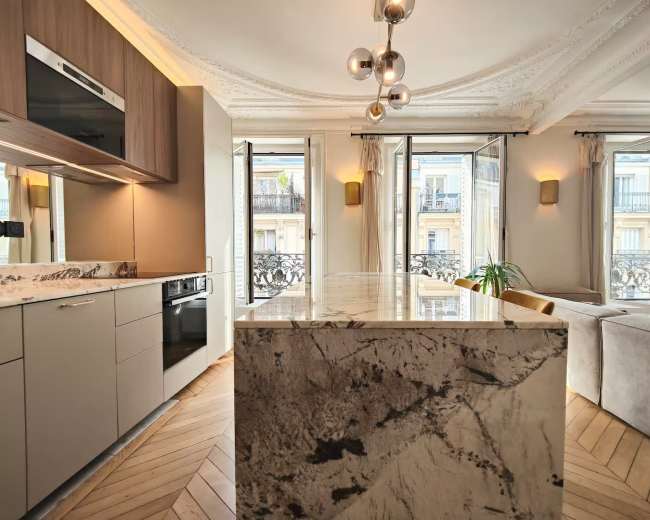 A modern kitchen features a marble island, sleek cabinetry, and large windows overlooking a balcony.