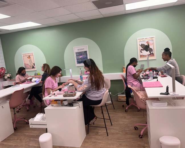 A group of five individuals are seated at manicure stations in a brightly lit salon, with green walls and fashionable posters in the background.