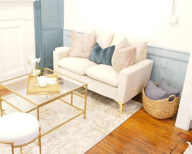 A light-colored sofa with decorative pillows sits across from a glass-top coffee table in a room with wooden flooring and a woven basket.