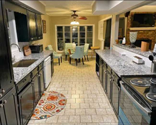 A modern kitchen features dark cabinetry, granite countertops, and a dining area with light-colored chairs, extending into a well-lit space.