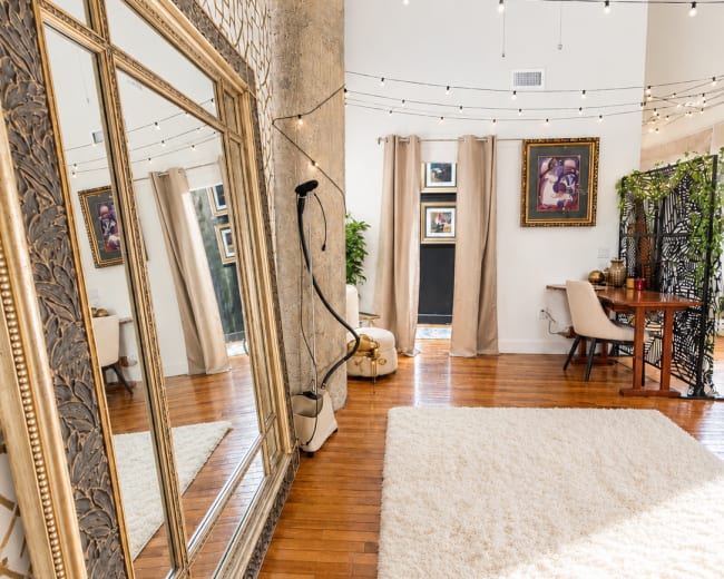 The image depicts a spacious room featuring a large ornate mirror, decorative string lights overhead, and a cozy seating area with a small desk and artwork on the walls.