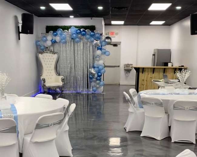 A decorated event hall with white tables, chairs, and a balloon arch in blue and silver behind a throne-like chair.