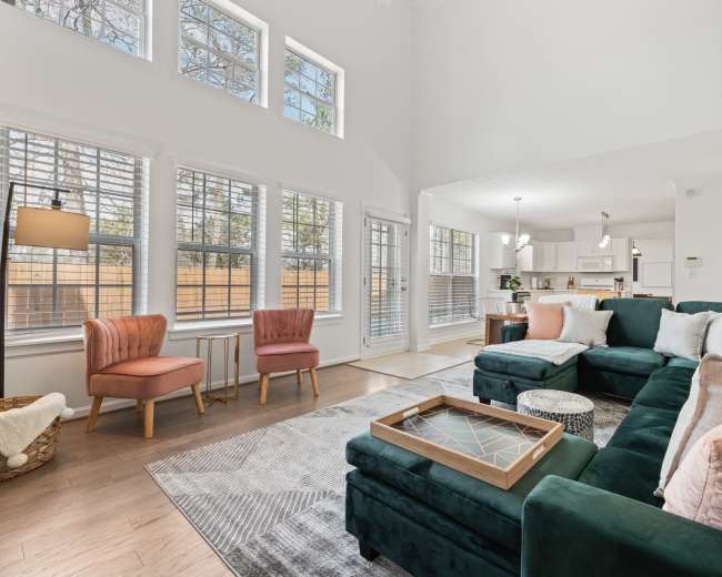 The living room features a green sectional sofa, two blush pink chairs, and large windows allowing natural light to fill the space.