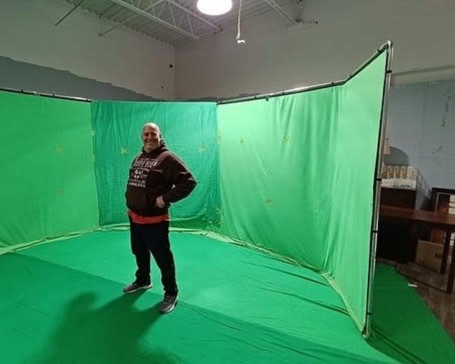 A man stands inside a green screen setup in a room with minimal furnishings.