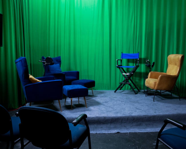 The image shows a studio setup with blue chairs, a yellow armchair, a director's chair, and a green backdrop.