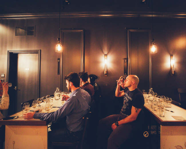 A group of people sit at a long wooden table in a dimly lit bar, each holding a glass and engaged in conversation.