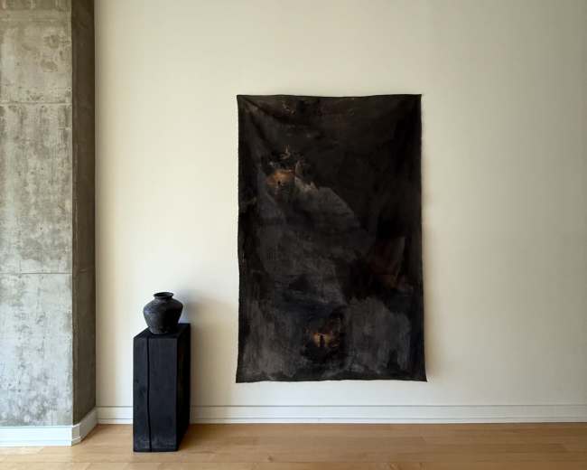 A dark textile is hung on a white wall above a black wooden pedestal with a ceramic vase on top, set against a backdrop of a concrete wall and wooden floor.