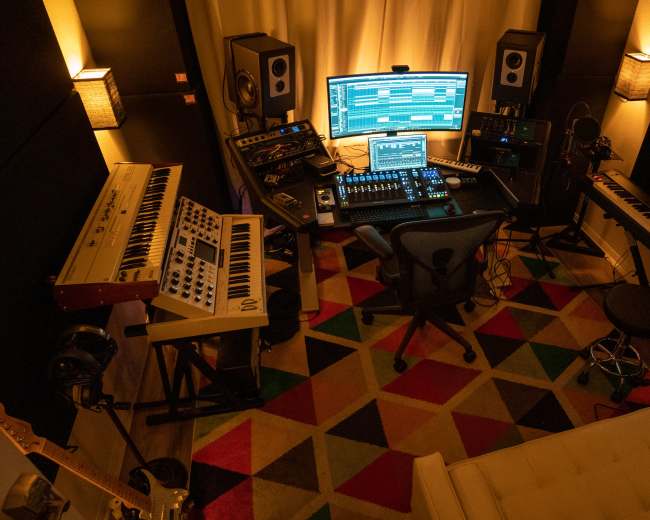 A music studio with multiple keyboards, a computer setup, and various audio equipment arranged around a central chair.