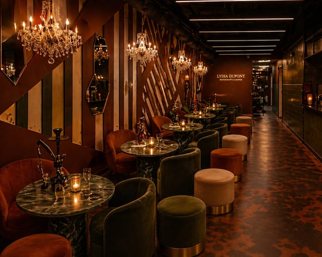 The image shows a stylish lounge with plush seating, decorative chandeliers, and a well-stocked bar in a contemporary interior design.