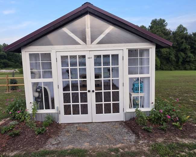 A small glass-enclosed building with double doors and a peaked roof is surrounded by flower beds and located on a grassy field.