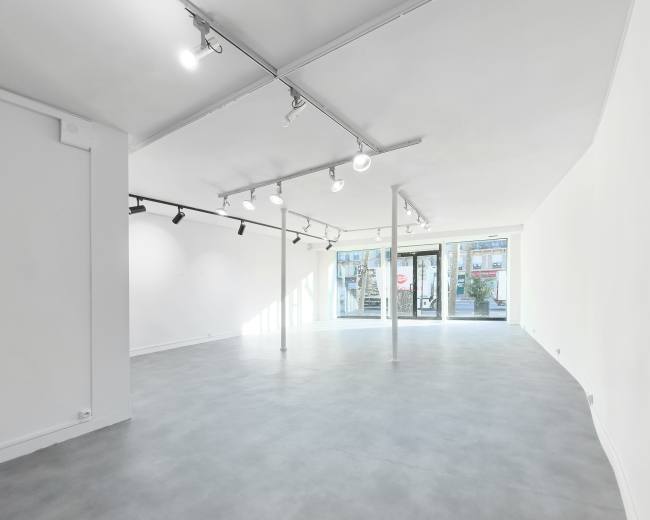 The image shows a spacious, empty room with a gray floor and white walls, featuring track lighting and large glass doors at one end.