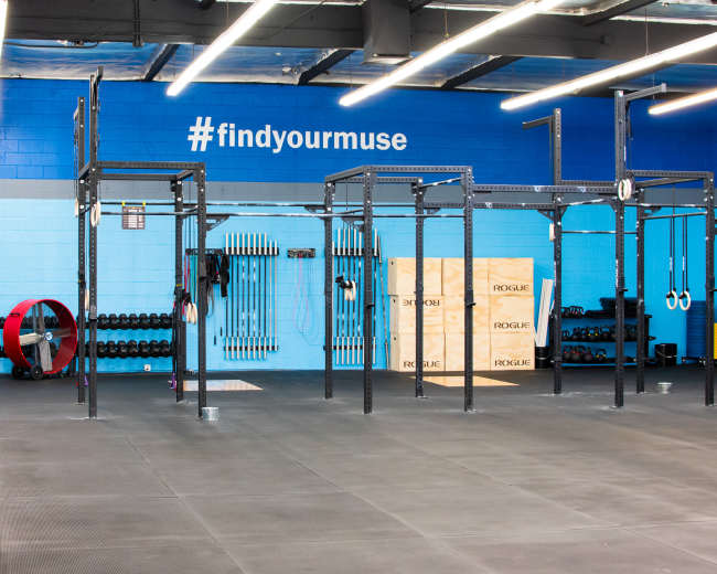 The image shows a spacious gym with weightlifting equipment, a pull-up station, and various fitness accessories against a blue wall marked with the hashtag "#findyourmuse."