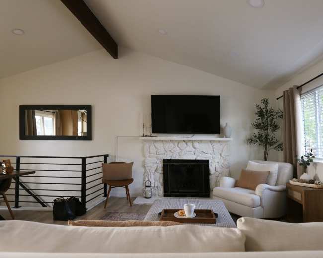 The image shows a cozy living room featuring a white stone fireplace, a mounted television, a comfortable armchair, and a small coffee table with a cup and snacks.