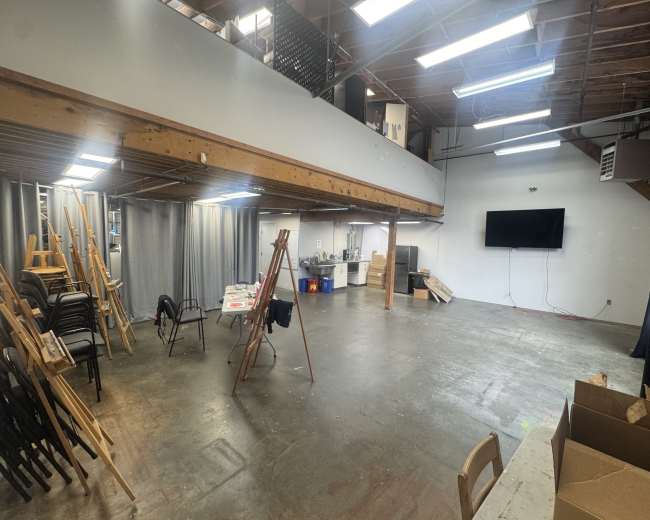 The image shows a spacious, partially illuminated studio or workspace with various chairs, easels, and a television mounted on the wall.