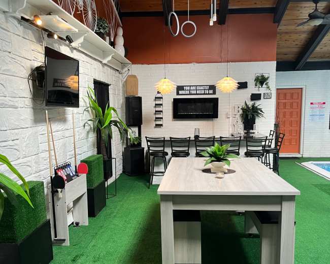 The image shows an indoor lounge area with a green carpet, a large dining table, chairs, plants, and wall-mounted televisions.