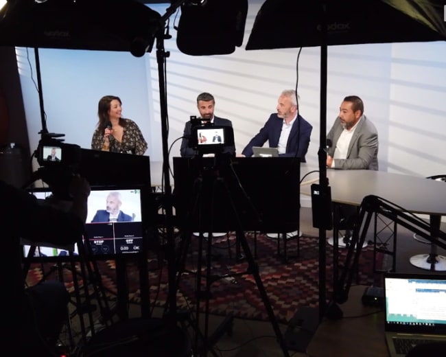 A group of four people participates in a discussion on a set equipped with cameras and lighting, while multiple screens display their conversation.