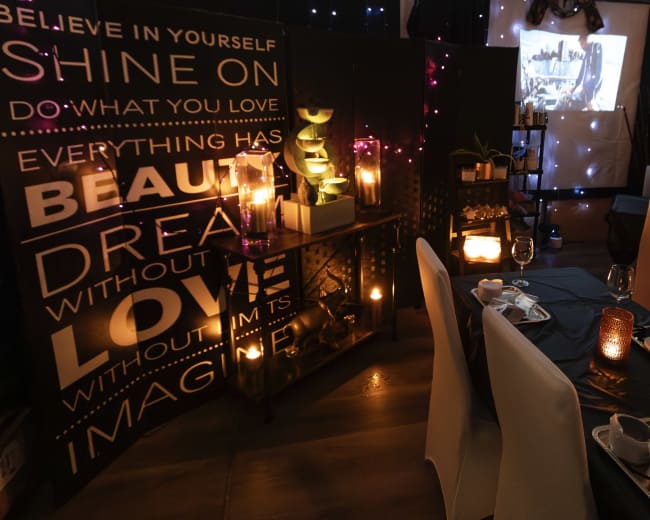 The image shows a dimly lit restaurant setting with tables arranged for dining, featuring decorative candles, a motivational wall display, and a projector displaying an image on a screen.