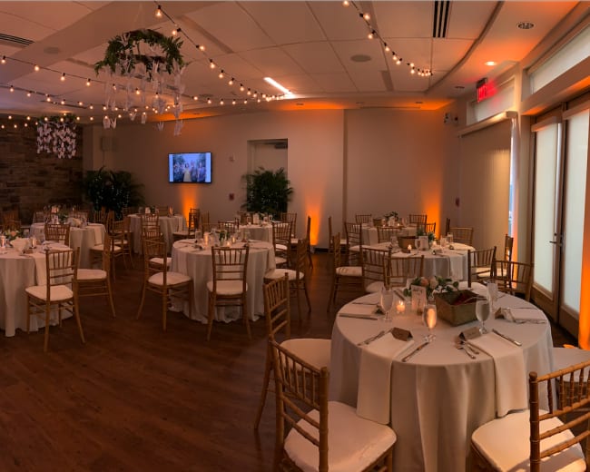 A well-decorated banquet hall with round tables set for an event, featuring a DJ booth and a large screen displaying photos.