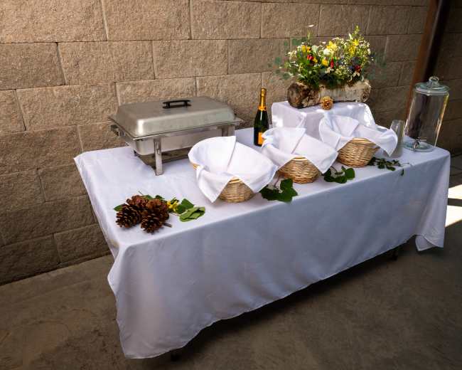 A buffet table is set up with four baskets lined with white napkins, a silver chafing dish, a decorative arrangement of flowers on wood, a bottle of champagne, and glasses.