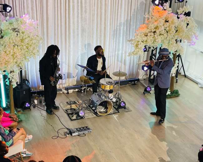 A small band performs onstage in front of an audience, surrounded by floral decorations and colorful stage lights.