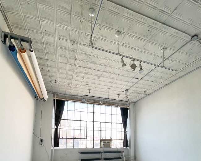 The image shows a large, empty studio space with white walls, a wooden floor, and a large window allowing natural light to enter.