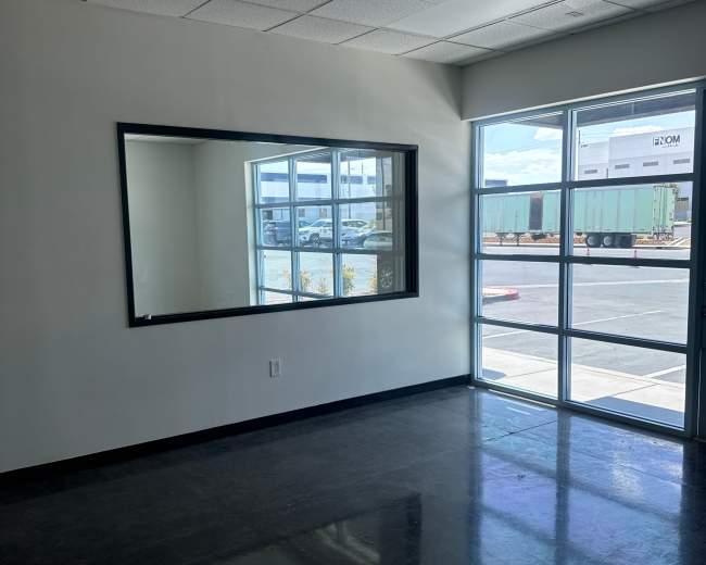 The image shows an empty room with a large mirror on one wall and a window that looks out onto a parking area with shipping containers.