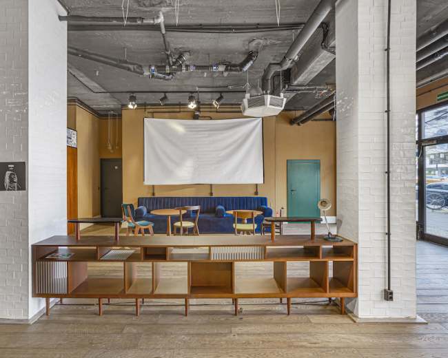 A modern interior space featuring a seating area with a blue couch, wooden tables, and chairs, alongside a large white projection screen and a wooden shelving unit.