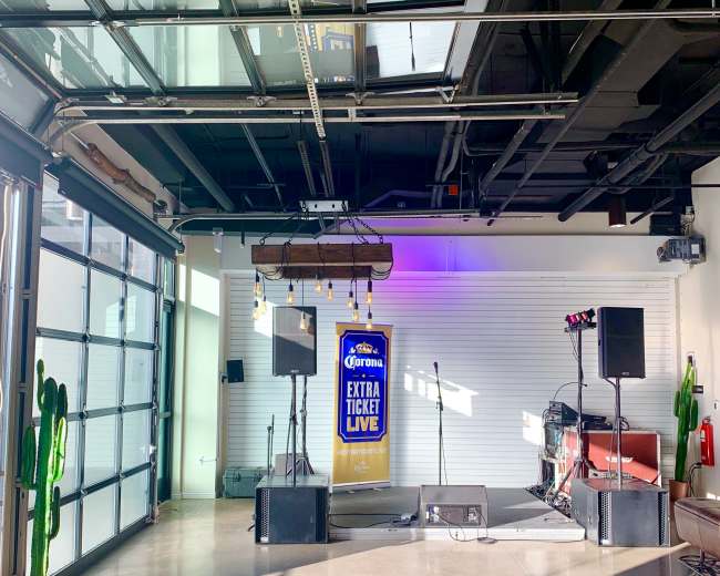 The scene shows a stage area in a modern indoor venue featuring a banner, microphones, and musical equipment, with large glass doors and potted cacti in the background.