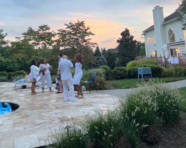 A group of people dressed in white gathers near a poolside as the sun sets behind a large house.