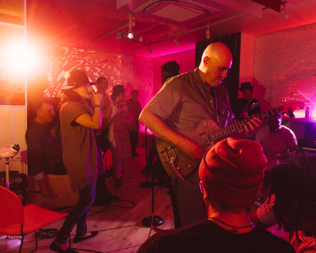 A musician plays guitar while people gather and enjoy a live music performance in a vibrant, illuminated room.