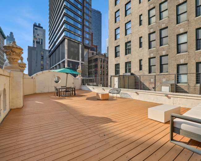 The image shows a spacious rooftop terrace featuring wooden decking, a seating area, and views of surrounding buildings in an urban setting.