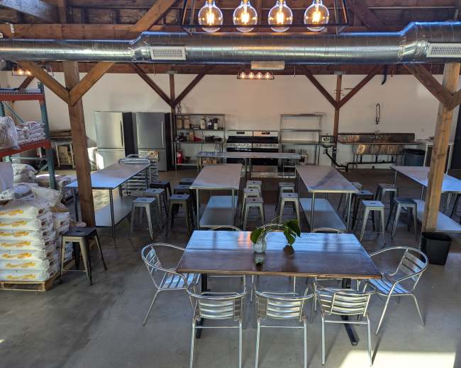 A spacious industrial kitchen setup with multiple stainless steel workstations, tables, and stools, illuminated by overhead lights.