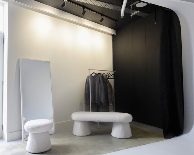 A minimalist dressing area featuring a full-length mirror, a clothing rack with gray garments, and two white, sculptural seating pieces.