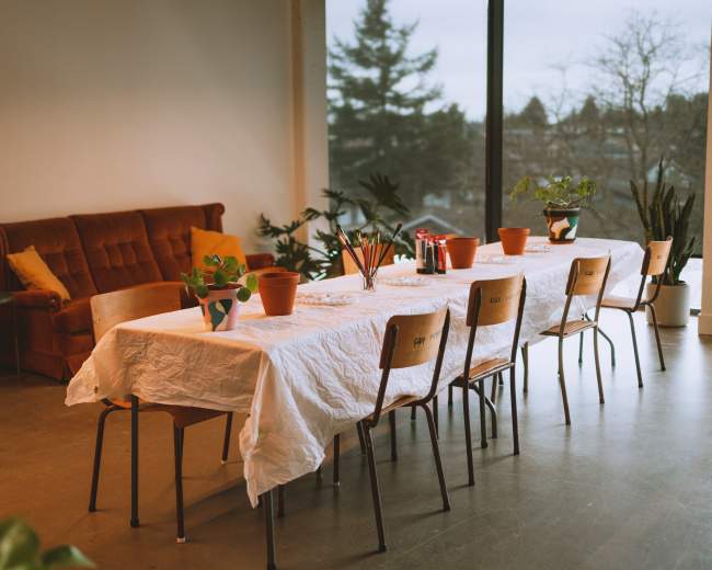 A long table covered with a white cloth is set up in a room with a sofa and potted plants, overlooking a window.