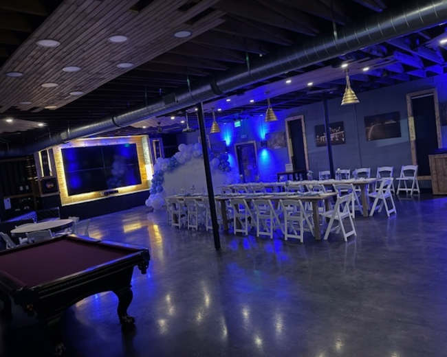 The image shows a spacious indoor venue with chairs and tables arranged for an event, featuring blue lighting, a large television screen, and decorative elements such as a balloon arch.