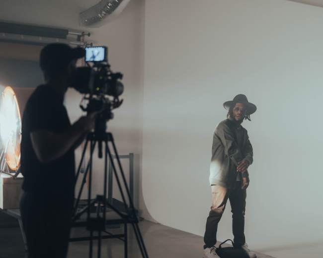 A person wearing a hat and sunglasses poses in front of a camera while a videographer captures the scene.