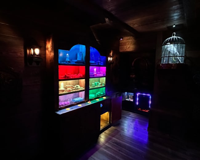 The image shows a dark room with a wooden display cabinet featuring shelves illuminated in various colors, alongside a decorative birdcage and dim lighting.