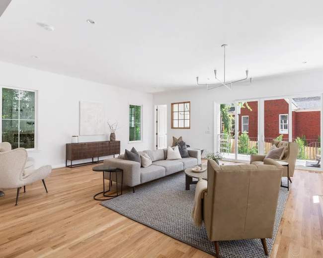 The living room features a modern layout with a large sectional sofa, two armchairs, a coffee table, and large windows that offer natural light and a view of the outdoors.