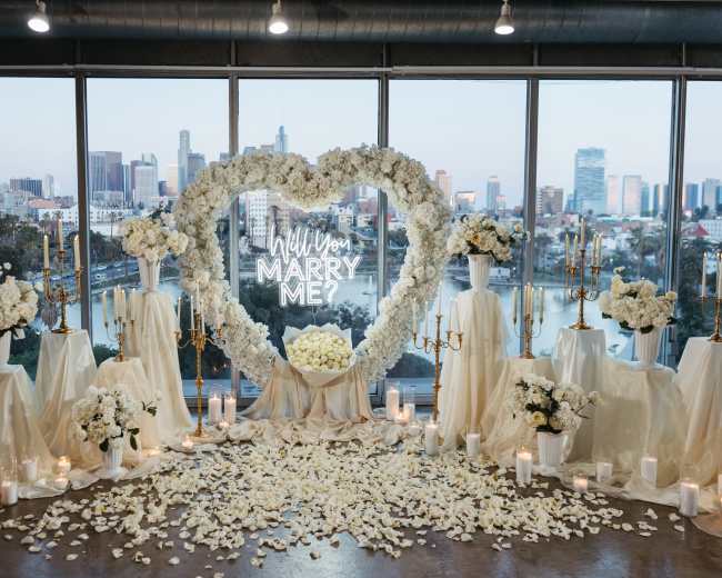The image shows a romantic proposal setup featuring a heart-shaped flower arrangement with a glowing "Will You Marry Me?" sign, surrounded by white flowers and candles, all set against a city skyline.