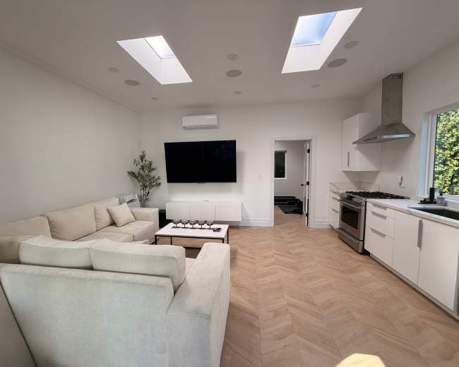 The image shows a modern living room with a white sofa, a large television mounted on the wall, and a kitchen area visible in the background.