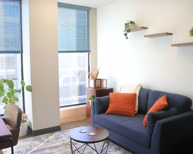 The image shows a modern office space featuring a dark blue sofa with orange pillows, a wooden desk with a chair, and a large window with blinds, complemented by small indoor plants and shelves on the wall.
